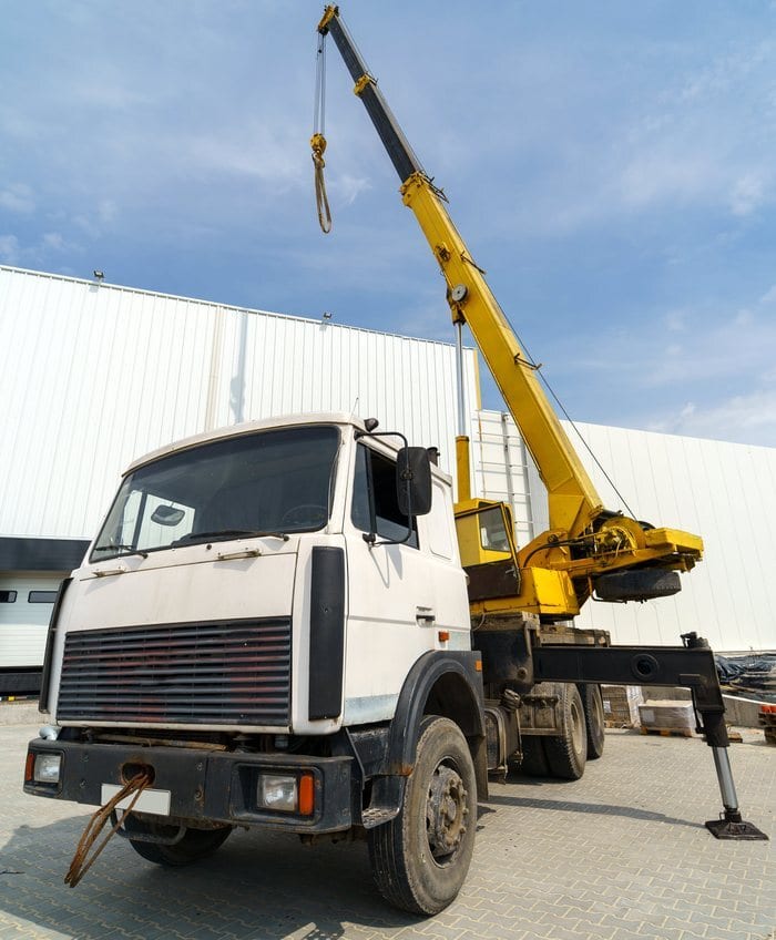 Truck mounted crane with its boom raised, parked against the background of a large warehouse